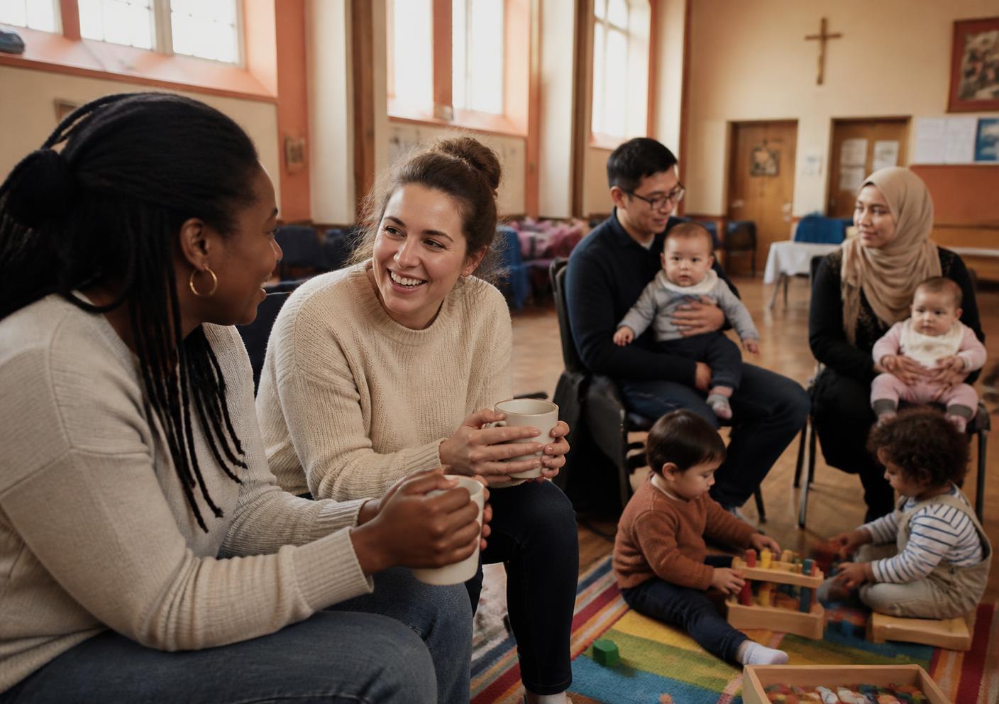 A diverse group of parents chatting over mugs of tea at a community baby group while their babies and toddlers play nearby